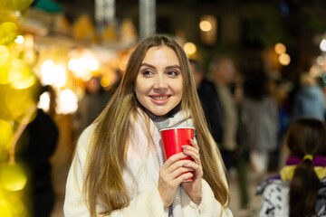 Happy girl walking in colorful Christmas street market, holding paper cup of warm drink