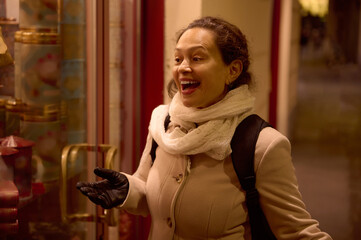 Happy smiling woman standing by illuminated showcase on city street, during Christmas fair in December at night.
