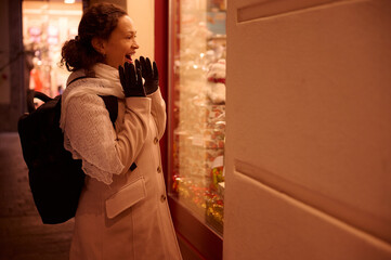 Happy cheerful woman smiles expressing amazement while standing at a gift shop window during a Christmas market at night