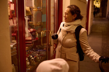 Pretty woman pushing baby pram, smiling and admiring the showcase at Christmas while walking along the street
