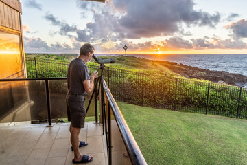 Mature Caucasian man standing on a lanai watching whales as the sun rising over the ocean,...