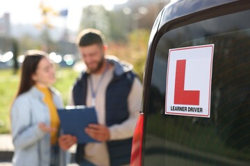 Learner driver and instructor with clipboard near car outdoors, selective focus on L-plate. Driving school - obrazy, fototapety, plakaty