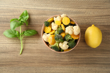 Frozen vegetables in bowl, basil and lemon on wooden table, flat lay