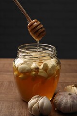Natural honey dripping from dipper into glass jar with garlic on wooden table