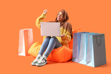 Young Muslim woman with laptop and shopping bags sitting on orange background