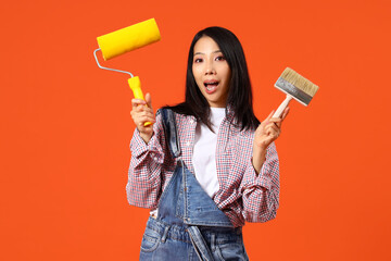 Young Asian woman with paint roller and brush on orange background