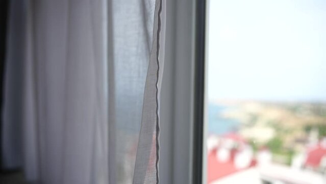 Sea view through the window with white transparent curtains slowly swaying in the wind. Resort, apartment with sea view. Leisure, relax, beauty, travel.