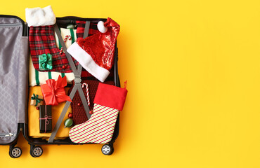 Open suitcase with clothes, Christmas gifts and decorations on yellow background