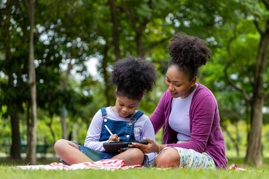 African American mother is teaching her young daughter to read using digital tablet while having a summer picnic in the public park for education and happiness