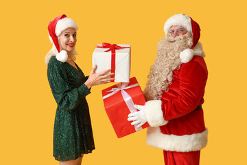 Santa Claus and beautiful mature woman with gift boxes on yellow background