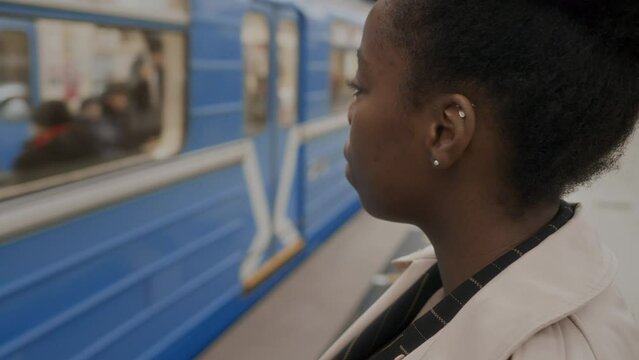 Medium closeup of young Black woman standing at subway platform close to railroad with train passing by