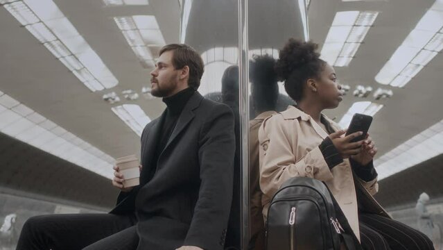 Low angle medium shot of ethnically diverse man and woman standing on different sides of stainless steel mirror column at subway station and waiting for train