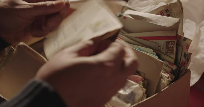 Person’s hands holding a  letter above  box full of old and yellowed letters and postcards. Close-up