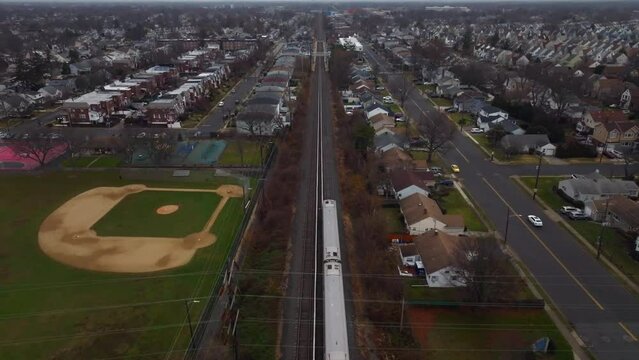 An aerial view of a Long Island railroad train traveling on a cloudy day. The camera dolly in over the tracks behind the train running through a residential neighborhood on Long Island, New York.