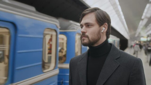 Waist up of Caucasian young bearded man standing at subway station filled with people and looking at moving train arriving at platform
