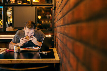 A man is enjoying his burger in a restaurant and watching videos on tablet.