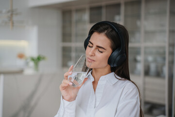 serene woman in white enjoys a moment of bliss with closed eyes, sipping water and listening to music on headphones, in a sleek kitchen.