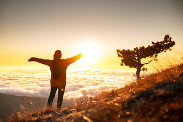 Happy young woman stands with open arms at mountain top above the clouds and looks at sunrise