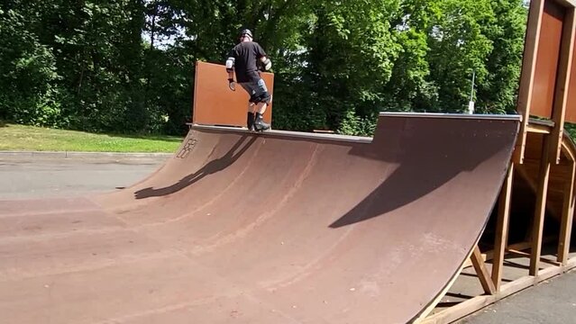 Ein Mann, der mit Inlineskates auf einer Halfpipe in einem Skatepark f&auml;hrt. Der Mann tr&auml;gt einen Helm und f&uuml;hrt einen Trick aus. Die Rampe ist aus Holz und gebogen.