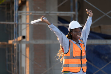 At the building site, a female engineer holds a blueprint in hand and raises her hands in delight. - obrazy, fototapety, plakaty