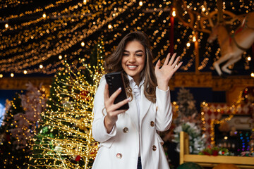 Young woman using smart phone video call talking with friend or boyfriend. Christmas tree decorated...