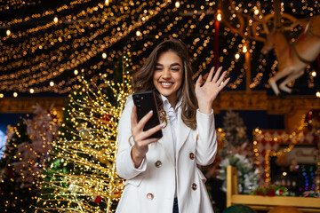 Young woman using smart phone video call talking with friend or boyfriend. Christmas tree decorated with ornament . Christmas and New Year holiday festival.