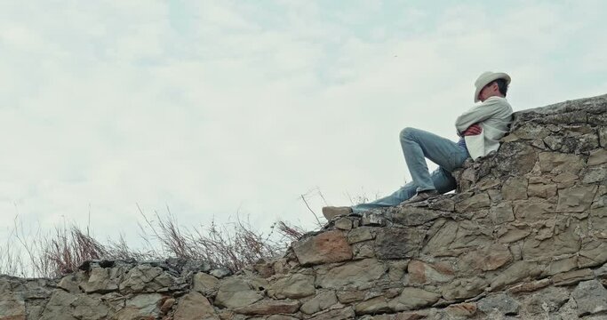 A young adult man in a hat sits on a medieval wall of wild stone against a blue sky and rests with his hat pulled down over his eyes.