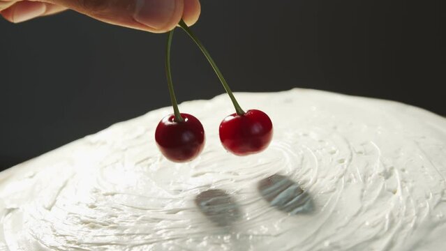Cherry on the cake. A hand puts two fused red cherries on a homemade white cake that rotates at an angle close up