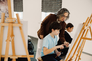 Teacher helping school boy to draw at easel during class in art school