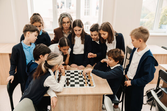 Group of kids and teacher watching chess game between two boys while standing around table in classroom - Powered by Adobe