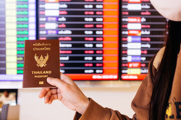 Asian woman wearing face mask and holding passport in front of flight information board