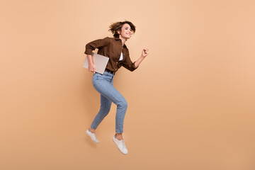 Fototapeta na wymiar Full length photo of pretty positive girl dressed brown shirt jumping high holding gadget empty space isolated beige color background