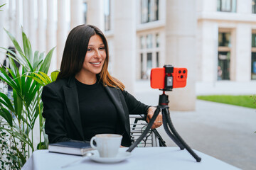 Confident female vlogger with a friendly smile, recording content using a smartphone on a tripod at...