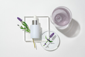 View from above of a white serum bottle on a glass podium, lavender flowers on a petri dish and a glass of water on a white backdrop. Blank labels for branding.