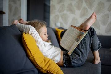 A little girl with blonde hair enjoys reading an interesting book in her house on the couch.