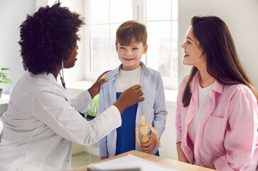 African American female doctor examining boy patient with stethoscope in medical office. Black...