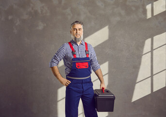 Portrait of a mature smiling bearded repairman holding a tool box in his hands on a gray wall...