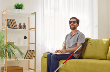 Happy independent disabled blind man in dark eyeglasses rests on sofa, holds Braille sign symbol...