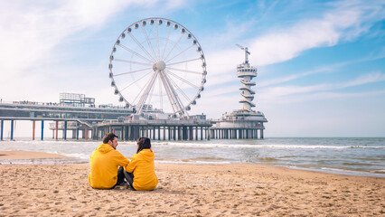 couple on the beach of Scheveningen Netherlands during Spring, The Ferris Wheel at The Pier at Scheveningen in the Netherlands, Sunny spring day at the beach  - obrazy, fototapety, plakaty