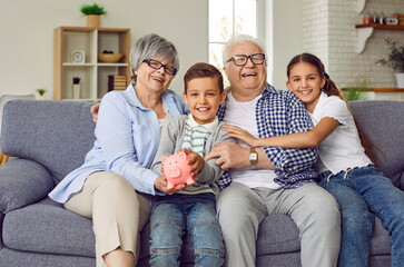 Portrait of a happy grandparents sitting on sofa with their grandchildren boy and girl in the living room at home looking at camera and holding piggy bank in hands. Saving money concept.