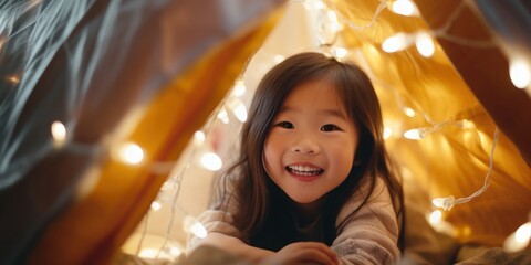 A young Asian girl is inside a blanket fort in a family room at night, softly lit by fairy lights hanging from the ceiling.