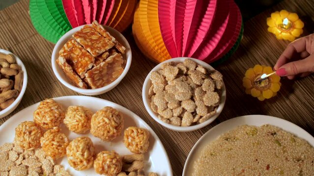 Pan shot of a woman's hand lighting up a flower-shaped candle marks the beginning of the Lohri festival with warmth and festivity. Table decorated with various sweets like Gajak  Peanuts Chikki  Re...