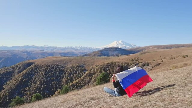 A young brunette woman with a developing Russian flag in her hands sits on the ground and looks at Mount Elbrus and the beauty of the mountains around. Back view