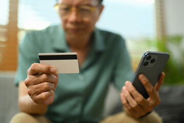 Closeup senior man holding credit card and smartphone paying for goods and online services.