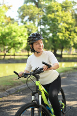 Fototapeta premium Portrait of happy retired woman in helmet riding bicycle in park on sunny summer day.