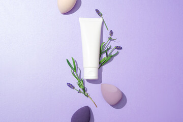 Top view of a white tube displayed on a purple background, surrounded by makeup sponges and lavender flowers. Mockup for advertising natural cosmetics with lavender extract.