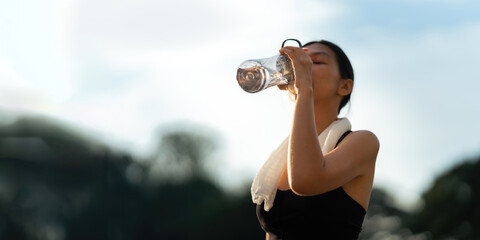 Beautiful young Asian woman stops to drinking water and wipe off her sweat after her evening run at park - obrazy, fototapety, plakaty