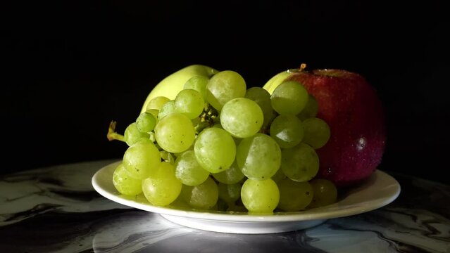 Two apples and a large bunch of ripe juicy green grapes rotate on a black background, illuminated by side light. Ideal for cooking, health and lifestyle content. 4k footage.