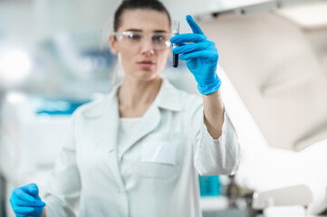 Fototapeta premium Young woman scientist working conducts research in a modern bio laboratory. The doctor holds a test tube with liquid in his hands and examines it