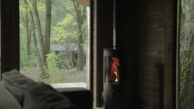 Cozy house chimney fire place with fire burning at the living room in tiny house, wooden wall on the background and beautiful window view to the green forest at summer, slow motion.